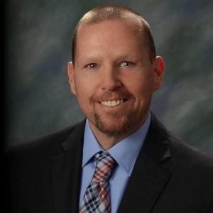 A picture of the City of Kaukauna Mayor, Tony Penterman wearing a black suit, blue shirt, and plaid tie