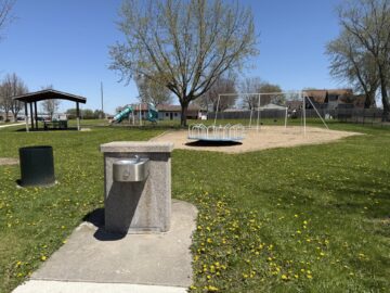 Picture of drinking fountain with swings, a pavilion, and green and tan playground equipment in the background