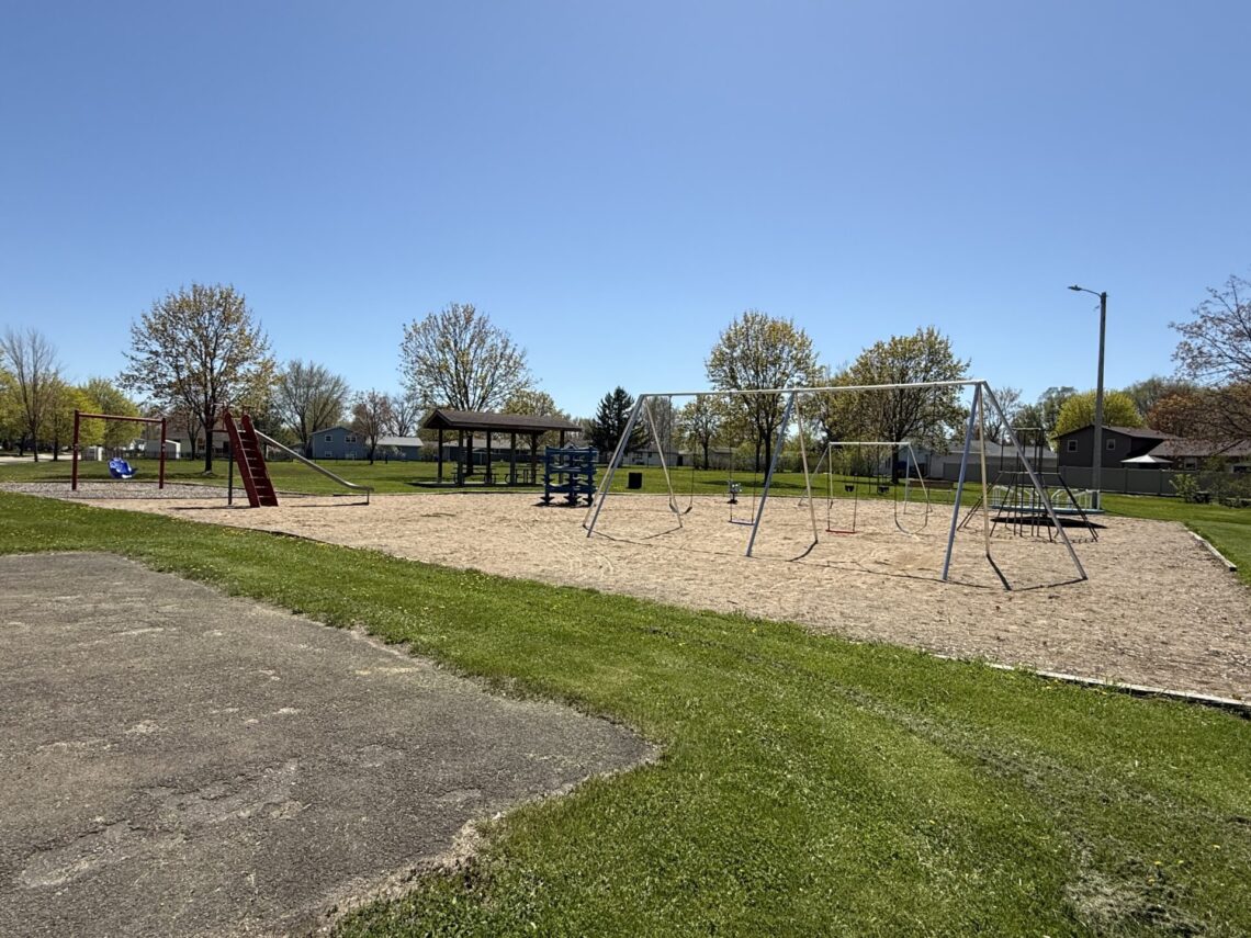 Picture of a swing set, climbing equipment and a metal slide and an open-air pavilion in the background