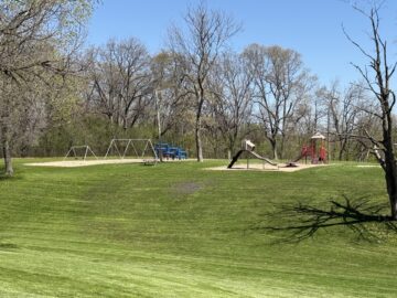 Picture of tan, red and blue playground equipment and swing sets on top of a grassy hill