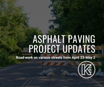 Image showing a road closure sign with additional signage about asphalt paving project updates by Kaukauna. The scene includes barricades and a forested area in the background. The project dates are from April 23 to May 2.