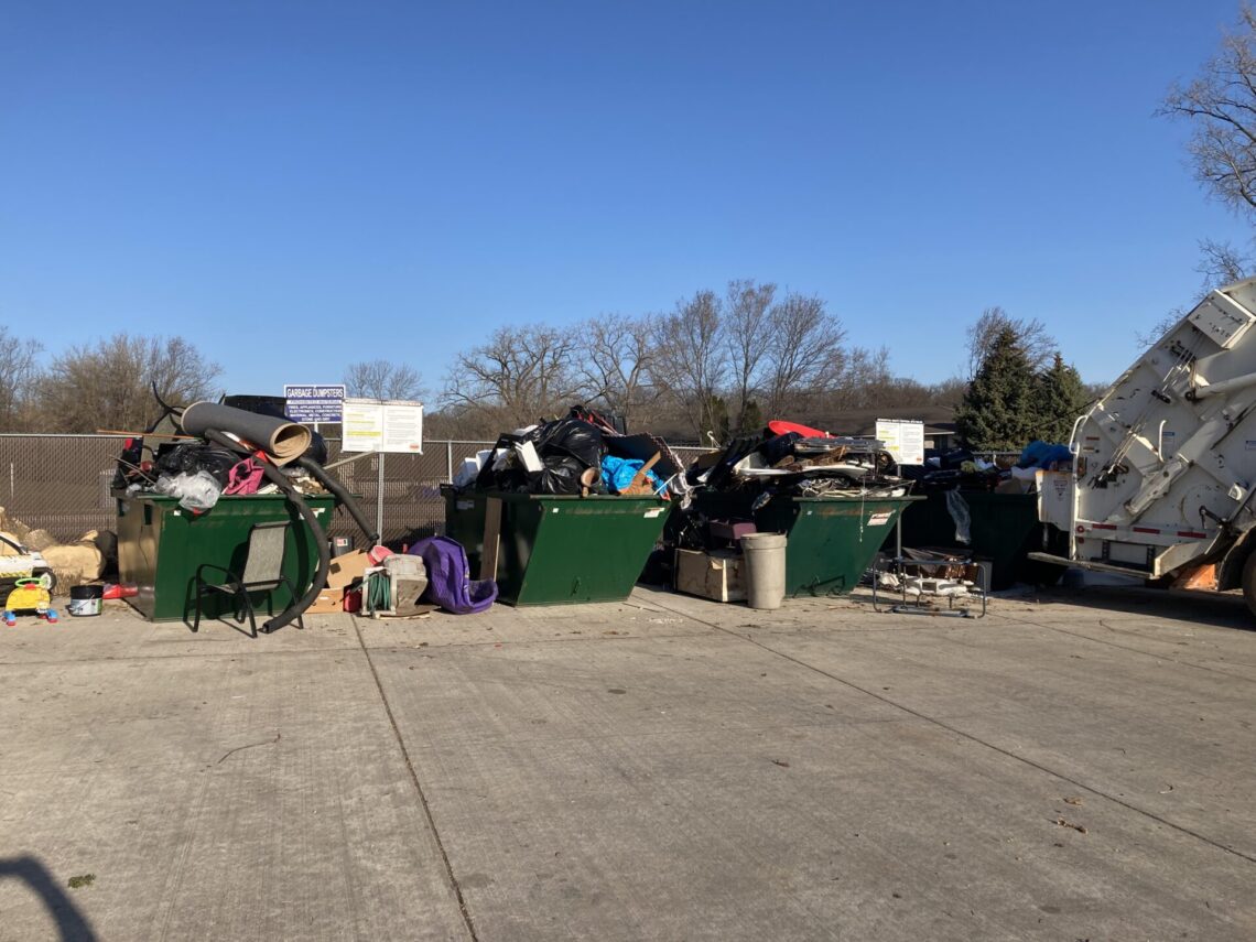 picture of three green dumpsters overflowing with carpet, wood, furniture, toys and other various items. Items are stacked alongside the dumpsters as well.