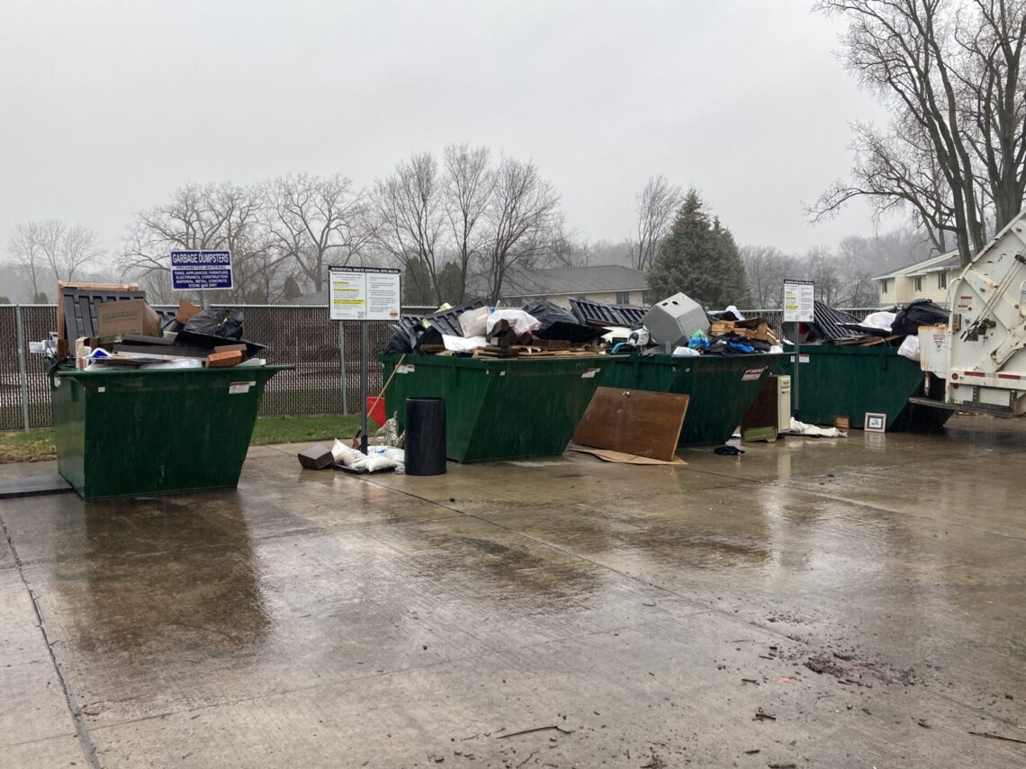 picture of four green dumpsters overflowing with carpet, wood, furniture, toys and other various items. Items are stacked alongside the dumpsters as well.