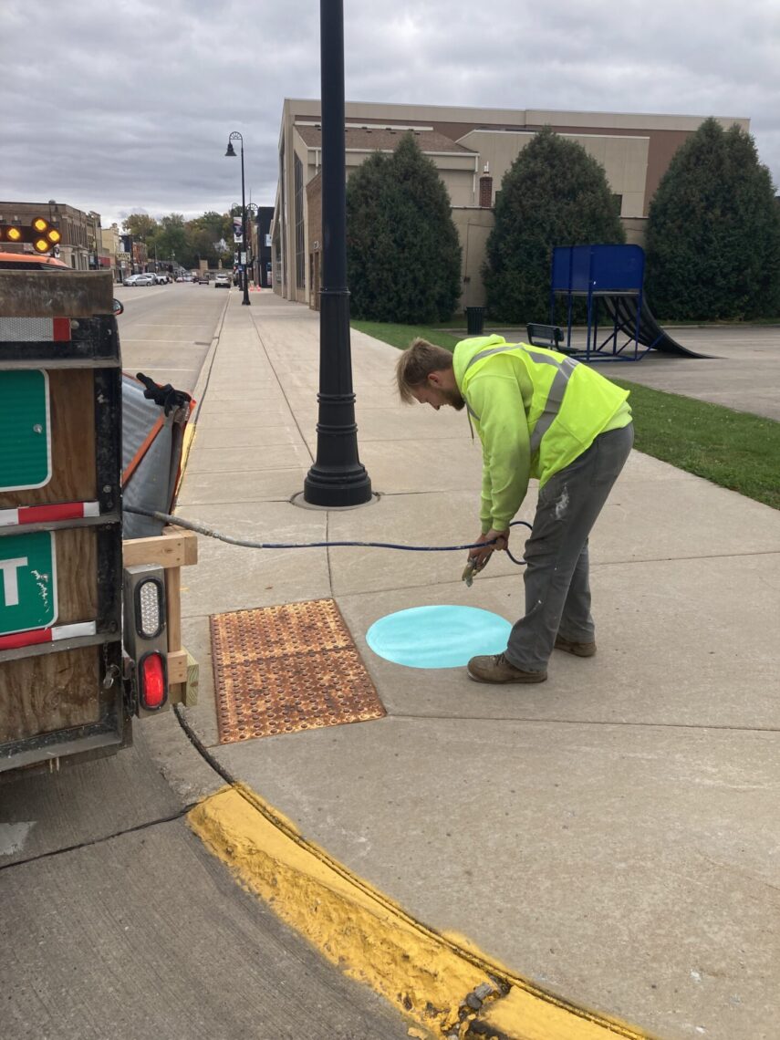A person wearing a bright safety jacket paints a blue circle on a sidewalk near a lamppost. The ground is marked with a textured orange ramp nearby. A trailer with traffic signs is positioned at the curb, and a large building is in the background.