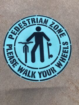 Blue circular sign on the ground with an icon of a person walking while pushing a bicycle and holding a skateboard. The text reads: 