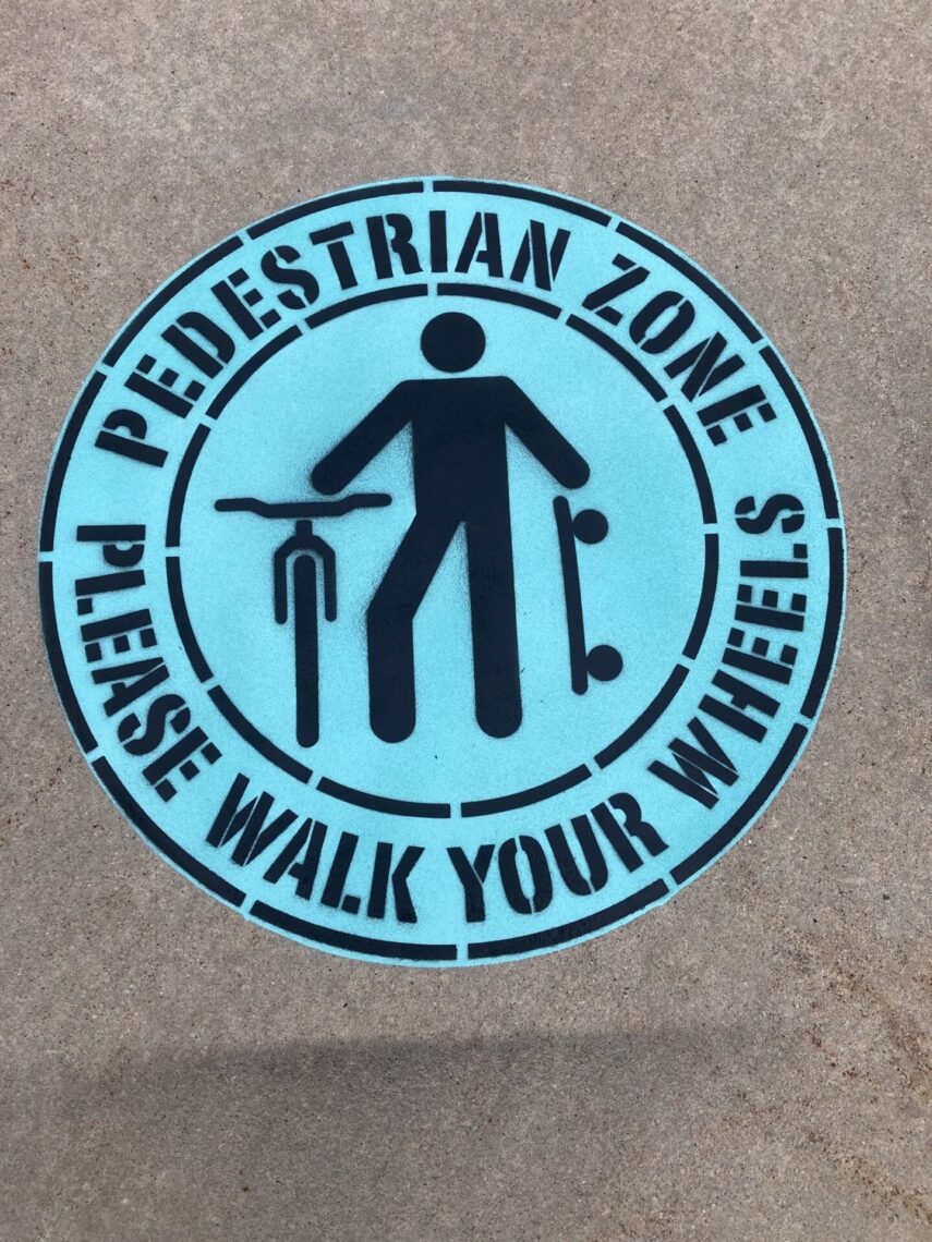 Blue circular sign on the ground with an icon of a person walking while pushing a bicycle and holding a skateboard. The text reads: 